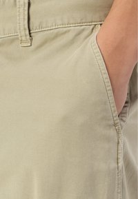 Tejido de algodón beige con una textura suave; cuenta con un bolsillo lateral y costuras cosidas, destacando su diseño a medida y detalles prácticos.