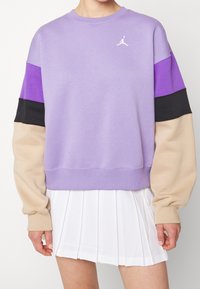 Sudadera morada con cuello redondo, mangas en contraste en negro y beige, y un logo blanco. Combinada con una falda blanca con pliegues.