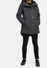 Personne portant une veste à capuche sombre, les mains dans les poches, des leggings noirs et des baskets noires, se tenant devant un fond uni.