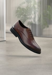Chaussure Oxford en cuir marron avec une finition lisse, six œillets, un bout arrondi et une semelle en caoutchouc noire, présentée en position flottante.