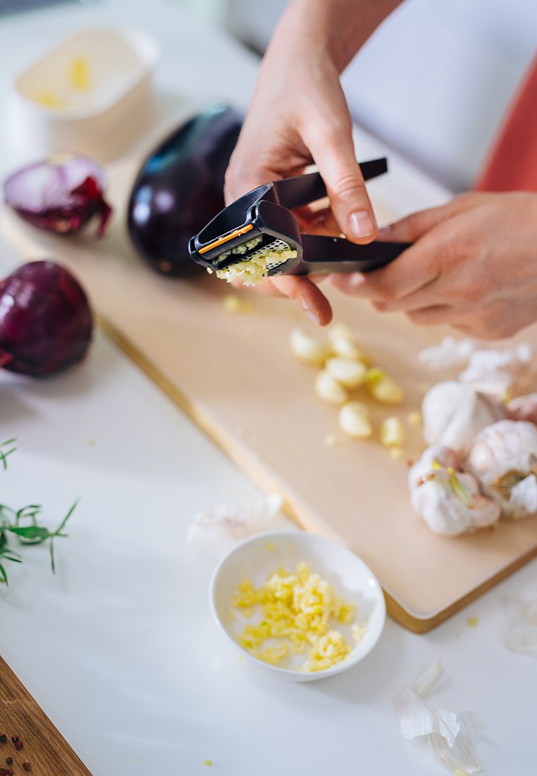 Schwarzer Knoblauchpresser in der Hand, gepresster Knoblauch wird auf ein Schneidebrett gedrückt. Rote Kohl und Knoblauchzehen in der Nähe, mit einer Schüssel voll gehacktem Knoblauch.