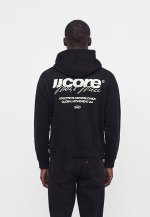 Mand iført sort hættetrøje med hvid tekst "Ucore Athletic Club Worldwide Global Movement Co. 1990" på ryggen, vendt væk fra kameraet.