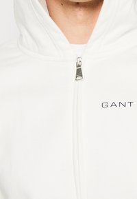 Sweat à capuche blanc zippé avec une fermeture éclair argentée et le logo "GANT" imprimé en noir sur la poitrine, porté par-dessus un sous-vêtement blanc.