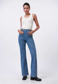 Τζιν παντελόνι σε μπλε χρώμα με καμπάνα και ψηλή μέση, με εμφανείς ραφές, συνδυασμένο με λευκό αμάνικο τοπ και μαύρα μποτάκια μέχρι τον αστράγαλο.