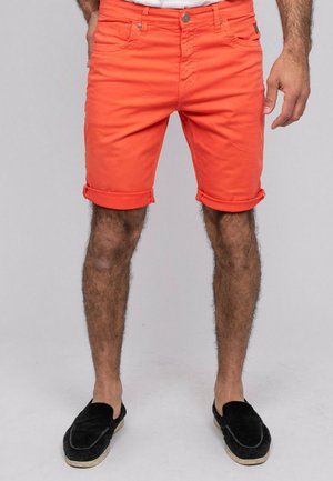 Homme portant un short orange vif retroussé et des chaussures noires sans lacets, debout les mains détendues le long du corps sur un fond gris.