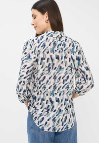 Langärmlige Bluse mit Bündchen, auf pinkem Hintergrund mit einem abstrakten blauen und türkisfarbenen Muster, Rundhalsausschnitt und geschwungenem Saum.