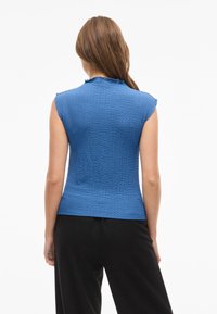 Blå, texturerad ärmlös topp med en volangkrage. Åtsittande design i kombination med svarta byxor, visad bakifrån.