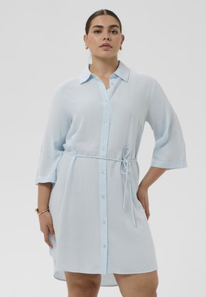 Vrouw die een lichtblauwe overhemdjurk met driekwartsmouwen draagt, geknoopt in de taille, staand tegen een effen achtergrond.