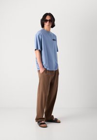 Tricou scurt de culoare albastră deschisă cu logo, asortat cu pantaloni bej largi și sandale negre, având un design cu vârf deschis.