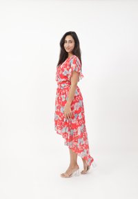 Robe fleurie avec un motif rouge et bleu, manches volantées et ourlet asymétrique. Associée à des sandales à talons transparents.