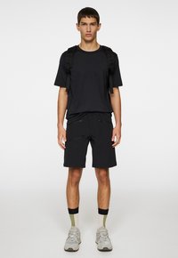 Svart sportt-shirt och matchande shorts med dragkedjor. Modellen har på sig grå sneakers och tvåfärgade strumpor. Enkel, funktionell design.