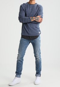 Blå långärmad skjorta med raglanärmar, lagd över en mörk t-shirt. Ljusblå jeans med slitna detaljer och vita sneakers.