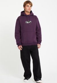 Paarse hoodie met een buidelzak, voorzien van een grafisch logo op de borst. Gecombineerd met zwarte wijde broek en sneakers.