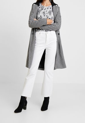 Frau trägt einen schwarz-weißen Hahnentritt-Mantel, graues Blumenhemd, weiße Hose und schwarze Stiefeletten, steht mit verschränkten Armen.