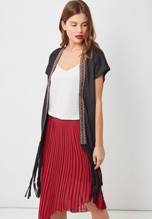 Mujer vestida con una blusa blanca, un chaleco negro de manga corta y abierto con un borde con estampado, y una falda roja plisada asimétrica, posando frente a un fondo blanco.