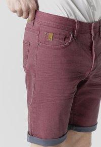 Hand zieht am Bund von hochgekrempelten, ausgewaschenen roten Jeansshorts, die über bloßen Beinen getragen werden, mit weißem Hemd, das hineingesteckt ist.