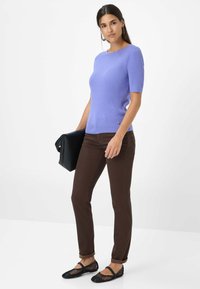 Lichtpaarse ribgebreide top met korte mouwen, gecombineerd met een bruine slim-fit broek en zwarte platte schoenen, terwijl ze een zwarte handtas vasthoudt.