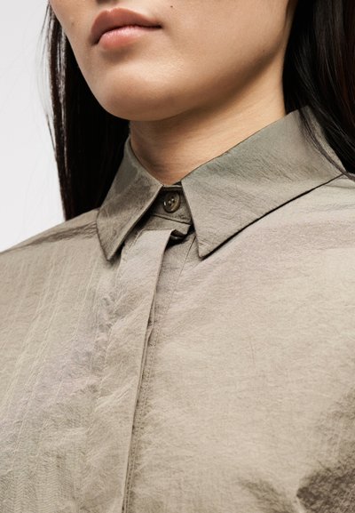 Κοντινό πλάνο ενός ατόμου που φοράει μπεζ πουκάμισο με κουμπιά και μυτερό γιακά, δείχνοντας το κάτω μέρος του προσώπου και την περιοχή του λαιμού.