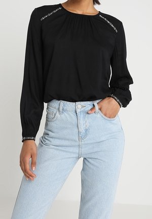 Vrouw met een zwart blouse met lange mouwen en zilveren versieringen op de schouders en manchetten, gecombineerd met lichtblauwe, hooggetailleerde jeans.