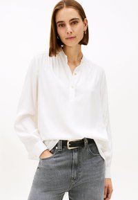 Blusa blanca de manga larga con cuello mandarín, botones y detalles plisados. Combinada con jeans grises de cintura alta y un cinturón negro.