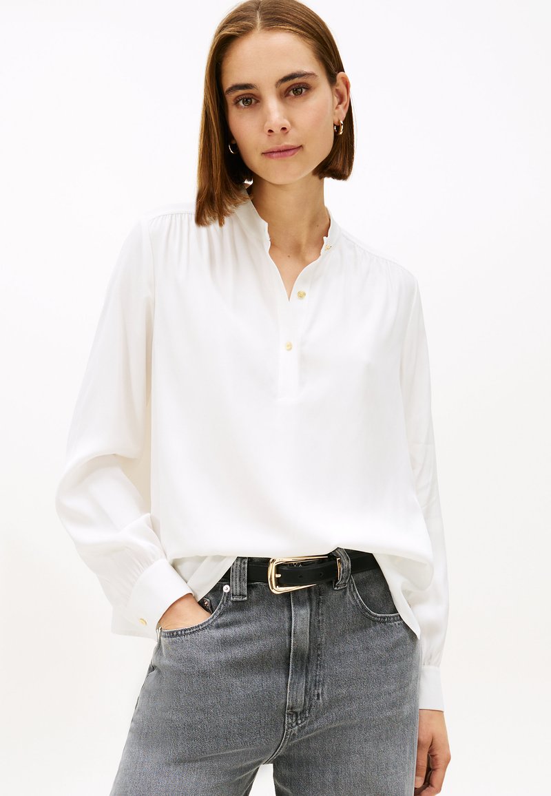 Blusa blanca de manga larga con cuello mandarín, botones y detalles plisados. Combinada con jeans grises de cintura alta y un cinturón negro.
