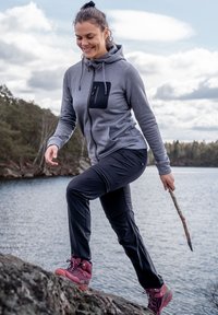 Grå zip-up hoodie med en svart bröstficka, svarta friluftsbukser och vinröda vandringsskor. Miljö nära en sjö med stenig terräng.