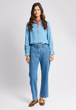Femme debout portant une chemise boutonnée bleu clair, un jean taille haute bleu, des ballerines marron et un foulard rouge autour du cou, sur un fond uni.