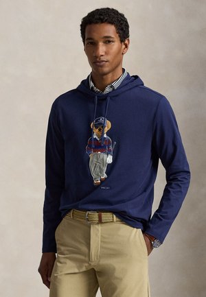 Uomo che indossa una felpa con cappuccio blu navy con grafica di un orso in abbigliamento da golf, camicia a righe sotto, pantaloni beige e cintura intrecciata, su sfondo semplice.