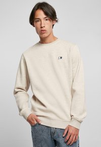 Sudadera beige de tejido suave, con cuello redondo y puños de canalé. Pequeño logotipo de estrella negra en el lado izquierdo del pecho. Combinada con vaqueros azules.