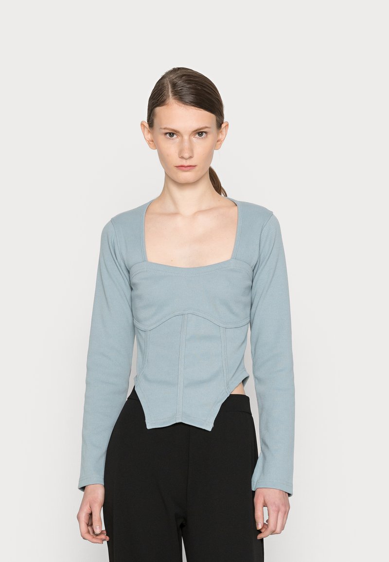 Camisola de manga longa azul claro com decote quadrado, textura canelada, design ajustado e costuras verticais. Usada com calças pretas.