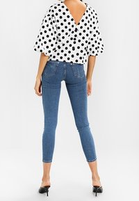 Weißes Top mit Pünktchen und tiefem V-Ausschnitt auf der Rückseite, kombiniert mit blauen Skinny Jeans. Die Jeans haben eine körpernahe Silhouette und dezente Gesäßtaschen.