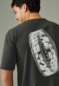 Tricou de culoare gri închis, confecționat din bumbac, având un grafic circular în alb și negru, cu text într-un aranjament distorsionat pe spate.