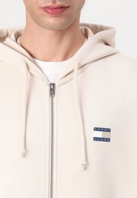 Cremfarvet zip-up hoodie lavet af blødt stof, prydet med et lille blåt og hvidt "TOMMY JEANS" logo patch på den øverste venstre bryst.