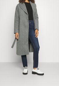 Donna che indossa un lungo cappotto a spina di pesce grigio, una maglia nera a coste, jeans skinny blu scuro e scarpe bianche con lacci e suola platform.