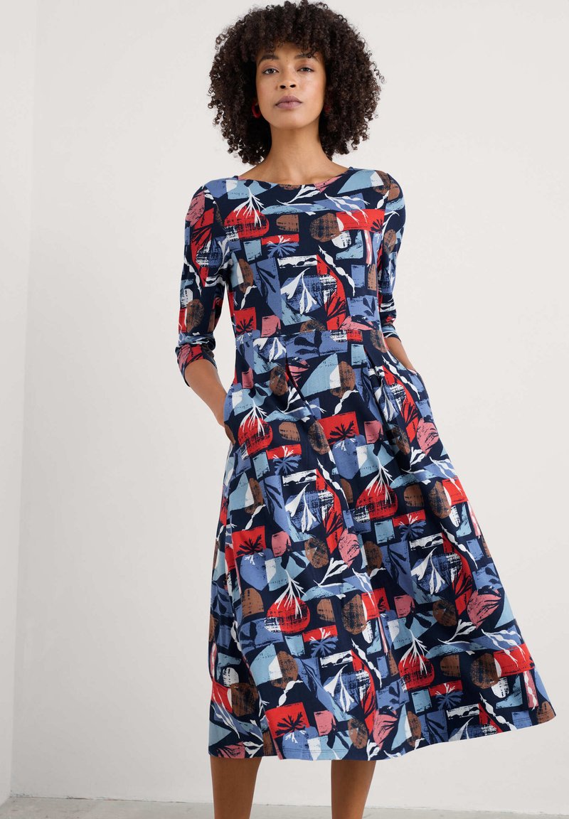Kes dress panjang sedang berwarna navy, menampilkan pola bunga abstrak yang cerah dalam warna merah, biru, dan coklat; memiliki kerah bulat dan dua saku samping.