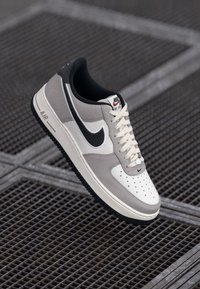 Nike Air Force 1 sportcipők világosszürke és krém színben, fekete Nike swoosh-sal, fehér fűzőkkel és perforált dizájnnal a lábujjrészen.
