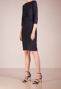 Robe de couleur bleu foncé, cintrée, avec des manches trois-quarts, un col bateau, et un tissu texturé. Associée à des sandales à talons noires avec des lanières.