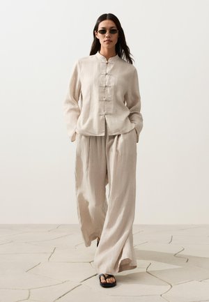 Femme marchant à l'intérieur portant un pantalon ample en lin beige, un haut à manches longues boutonné assorti, des sandales noires et de petites lunettes de soleil foncées.