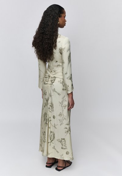 Femme aux longs cheveux bouclés portant une longue robe crème à motifs floraux et esquissés avec des sandales noires à brides, debout devant un fond uni.