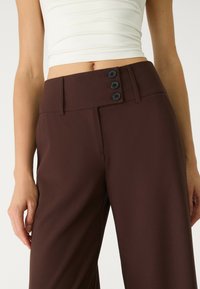 Bruna högmidjade byxor med en mjuk textur, utrustade med tre svarta knappar vid midjan och sidofickor.