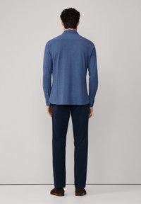 Un hombre se encuentra de pie frente a una pared gris clara, vistiendo una camisa de manga larga azul, pantalones oscuros y zapatos marrones.