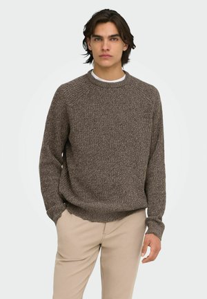 Jeune homme aux cheveux foncés portant un pull en tricot marron et un pantalon beige, debout avec une main dans la poche sur un fond clair.
