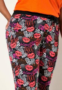 Mønstrede joggers med en blanding af røde læber, blomsterdesigns og grafisk tekst på en sort baggrund. Elastisk talje og sidelommer.