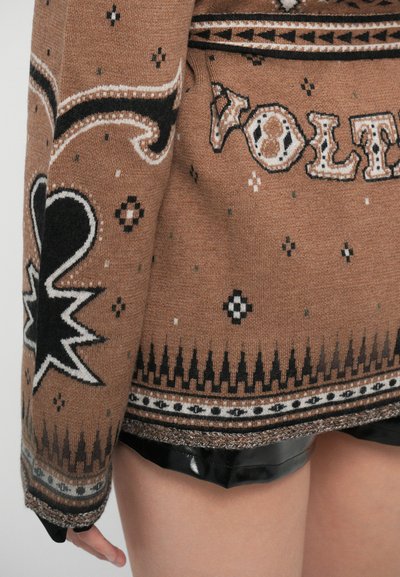 Pull en tricot marron avec des motifs noirs et blancs, incluant des formes géométriques et du texte. Ourlet côtelé et détails en tissu texturé.