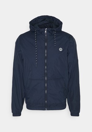 Veste coupe-vent à capuche bleu marine avec fermeture éclair à l'avant, poignets élastiques, cordons de serrage et deux poches avant, présentée sur un fond blanc.