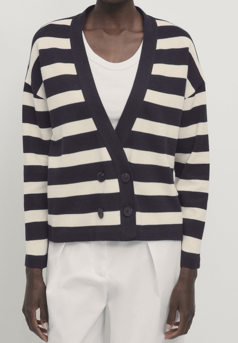 Prążkowany kardigan w kolorach granatowym i kremowym, z dekoltem w kształcie litery V, dwoma guziczkami z przodu oraz luźnym krojem i długimi rękawami. Gładka dzianinowa faktura.