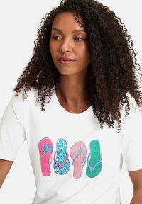 T-shirt blanc avec un graphique de quatre tongs en rose, bleu et vert, ornées de motifs et de détails. Coupe décontractée et manches courtes.