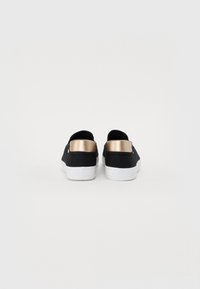 Svarta canvas slip-on skor med vit sula och metallisk guldaccent på hälen, med en mjuk textur och minimalistisk design.