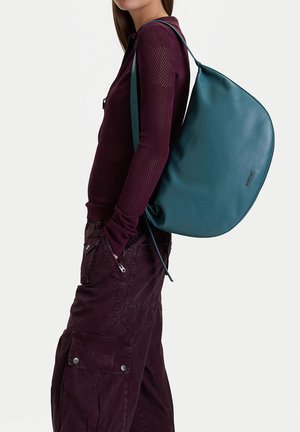 Mujer con pantalones cargo burdeos y top de manga larga de malla, llevando un bolso grande de cuero azul verdoso al hombro.