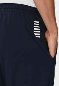 Pantaloni sportivi blu realizzati in tessuto morbido, con vita elastica, tasca laterale e strisce verticali bianche sulla tasca.
